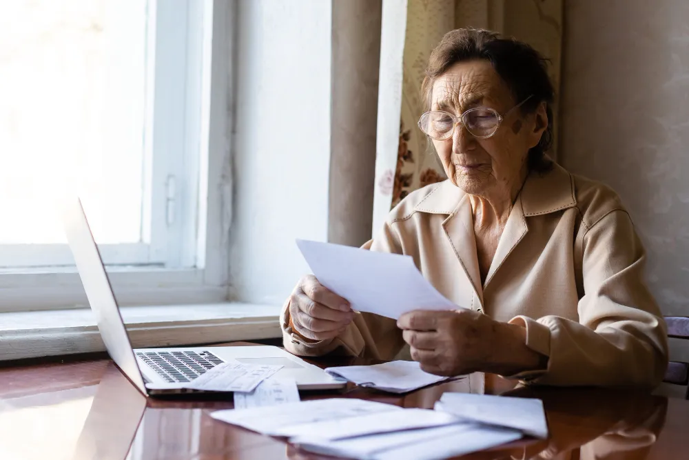 Idosa com óculos analisa documentos perto de laptop, buscando informações sobre BPC para idoso acima de 65 anos.